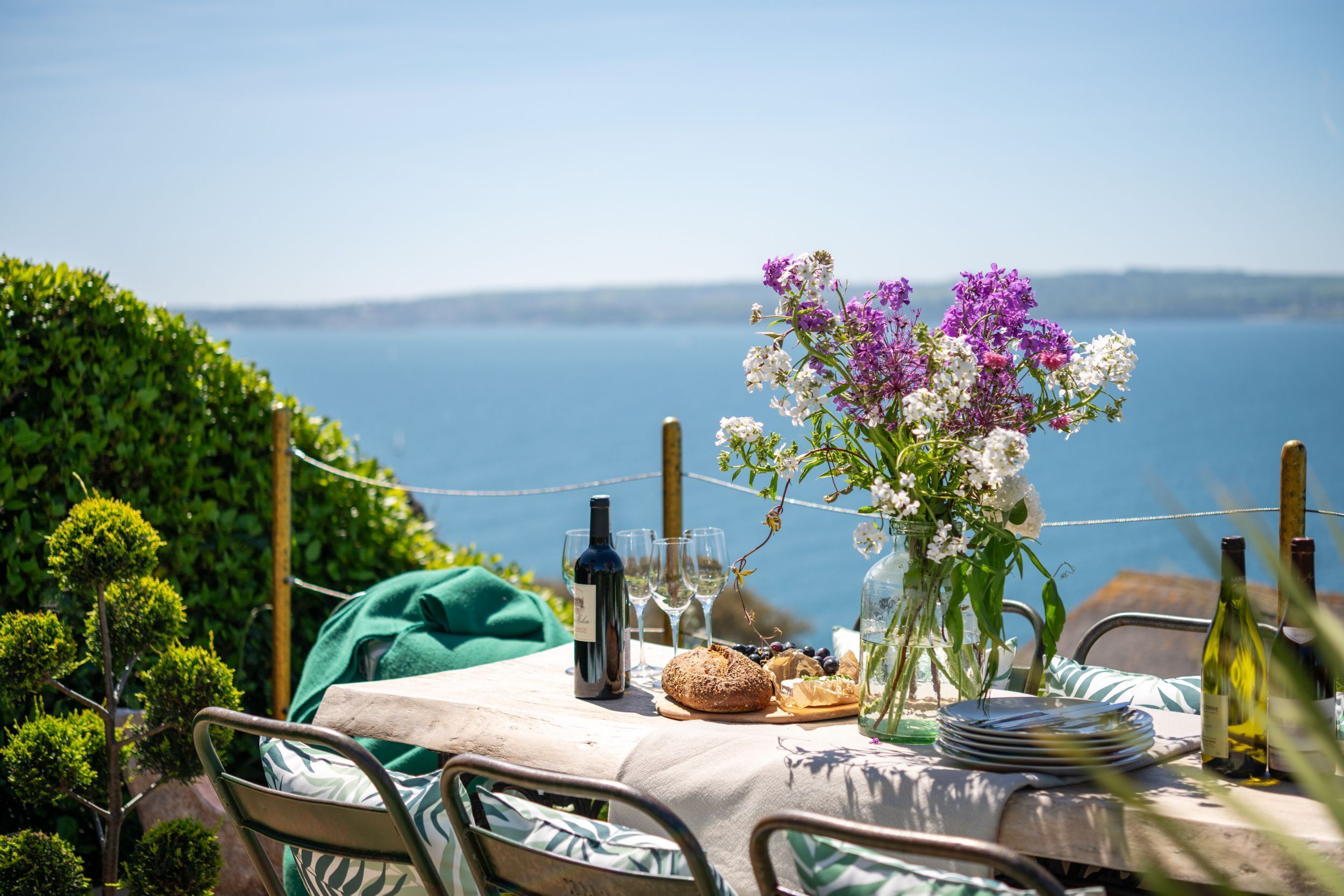 A table set with wine glasses, a bottle of wine, a loaf of bread, and a vase of flowers, overlooking a body of water.