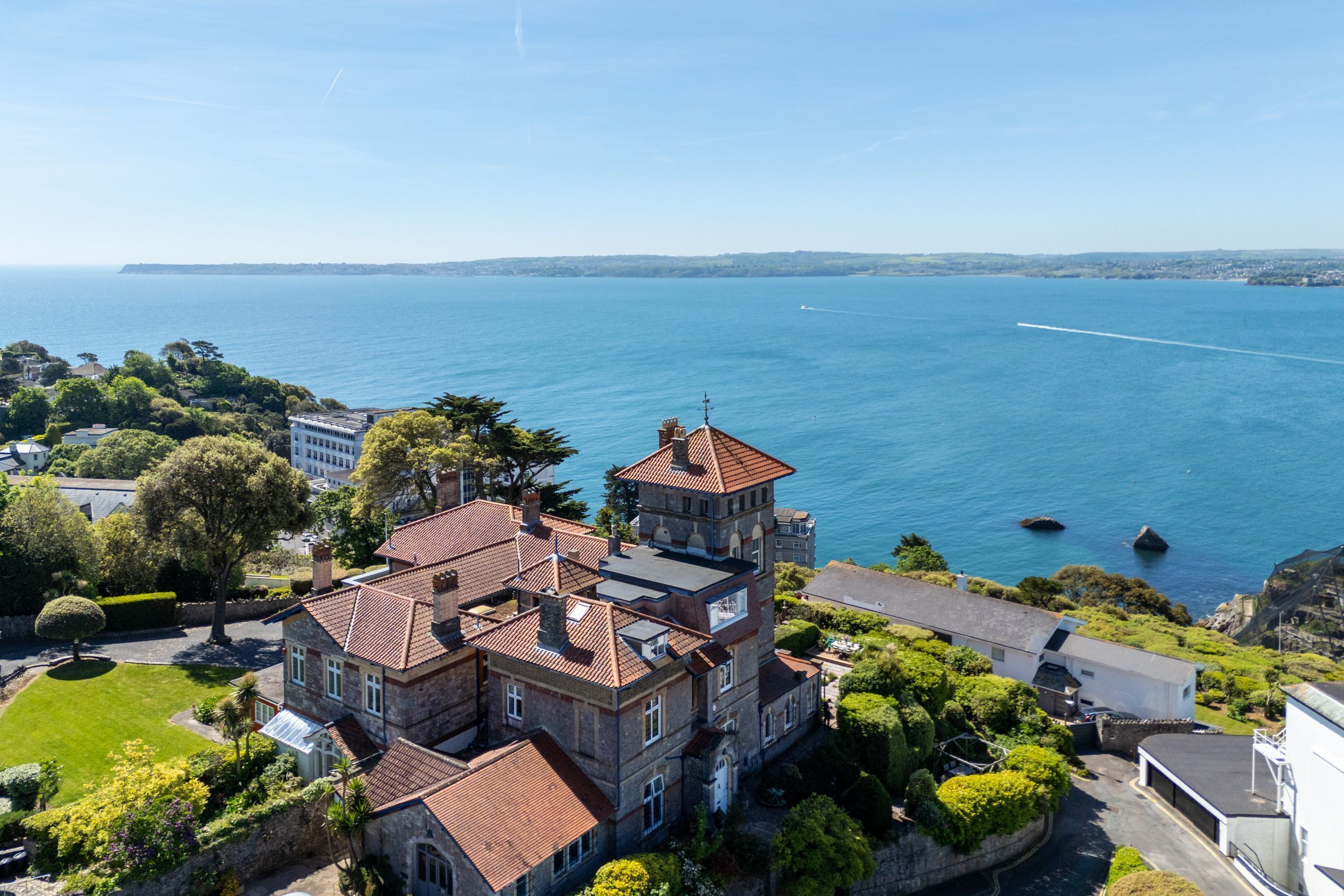 A large, multi-story stone building with a red-tiled roof, surrounded by lush greenery and overlooking a large body of water.