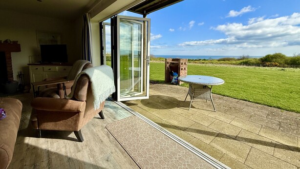 A living room with a sofa, a TV, and a glass door leading to a patio with a table and a brick structure. - Len's Lookout -  a cottage that sleeps 8 guests  in 4 bedrooms (Filey)