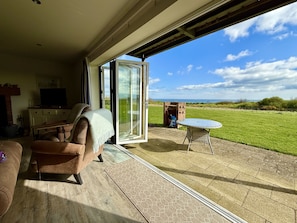 A living room with a sofa, a TV, and a glass door leading to a patio with a table and a brick structure. - Len's Lookout - a cottage that sleeps 8 guests in 4 bedrooms (Filey)