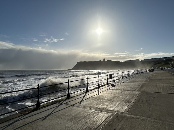 A coastal walkway with a railing, waves, and a distant hill with structures. - Len's Lookout - a cottage that sleeps 8 guests in 4 bedrooms (Filey)