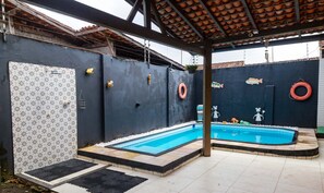 Une piscine avec une terrasse carrelée, un espace couvert doté d’un toit carrelé et un mur orné de carreaux décoratifs et de bouées de sauvetage.