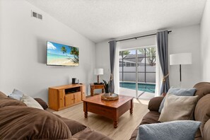 Sala de estar com vista para a piscina, mesa de centro de madeira, televisão e sofá.