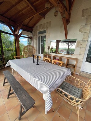 A covered patio with a table set for dining, wicker chairs, a bench, and a view of the outdoors.