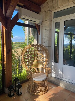 Une chaise en osier avec un coussin, posée sur un sol carrelé, près d’une fenêtre donnant sur un paysage verdoyant.