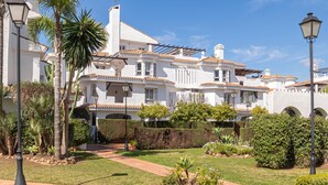 A multi-story building with a white facade, terracotta roof tiles, and a landscaped garden with palm trees and shrubs.