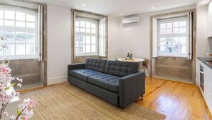 Een moderne woonkamer met een grijze bank, grote ramen en een keukenhoek.