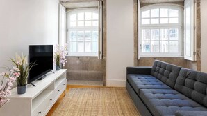 Een moderne woonkamer met een flatscreen televisie op een witte console, een grijze bank en twee ramen met witte kozijnen.