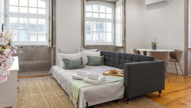 Een moderne slaapkamer met een slaapbank, twee ramen, een klein tafeltje met een plant en uitzicht op de stad.