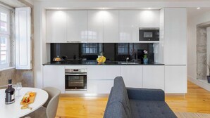 Een moderne keuken met witte kasten, een zwart aanrecht en roestvrijstalen apparatuur. Er is een eethoek met een tafel en stoelen, en een grijze bank in de woonkamer.