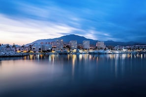 A coastal cityscape at dusk with buildings reflecting on calm waters, a marina with boats, and a mountain in the background. - Oceana Breeze Apartment with Pool View (Estepona)