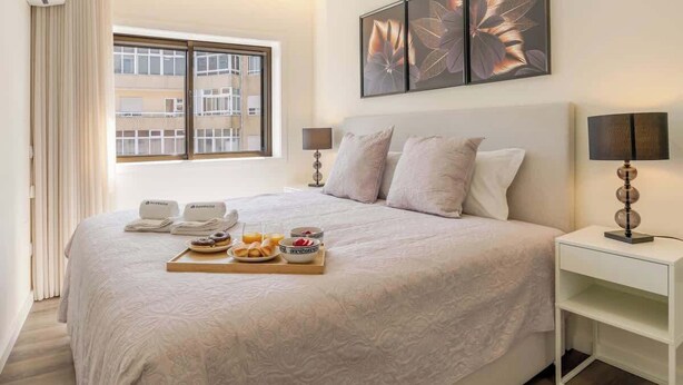 A neatly made bed with a tray of breakfast items, a window with blinds, and a wall with floral artwork.