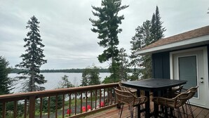 A wooden deck with a table and chairs, overlooking a lake with trees and a building in the background. - Comfort and Convenience. Family and pet friendly. Close to the action! (International Falls)