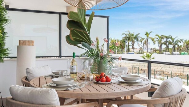 A round wooden table with white plates, glasses, and a bottle of wine, surrounded by wicker chairs and a large plant. - Lujo Apartments Estepona (Estepona)