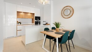 Eine moderne Küche mit weißen Schränken, einer zentralen Kochinsel und einem Essbereich mit Holztisch und blaugrünen Stühlen.