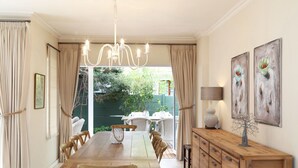 A dining area with a wooden table and chairs, a chandelier, and a view of the garden through open curtains.
