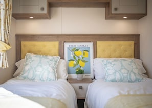 A hotel room with two beds, each with a yellow headboard, white bedding, and patterned pillows. A poster titled 'FRUIT MARKET' hangs between the beds.
