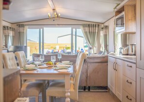 Un espace repas avec une table dressée pour quatre personnes, un lustre et une vue dégagée sur l’extérieur grâce à de grandes fenêtres.