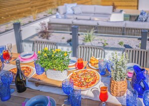 Un espace de restauration en plein air avec une table en bois dressée pour un repas, entourée de chaises et d’un canapé.
