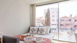 A hotel room with a bed, pillows, a tray of fruit, towels, and a view of buildings through a large window.