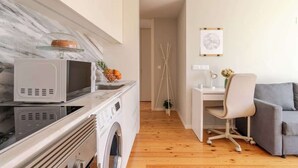 A modern kitchen with a microwave, oven, and washing machine. There is a dining area with a desk, chair, and a grey sofa.
