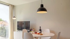 A dining area with a round table, white chairs, a black pendant light, and a TV on a white cabinet.