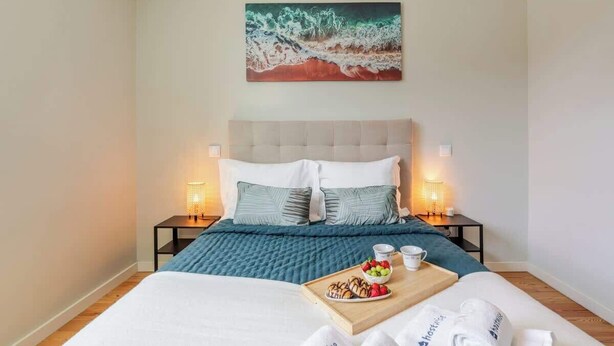 A neatly made bed with a blue blanket, two pillows, and a tray with fruits and a cup. There is a painting above the bed and two bedside lamps on either side.