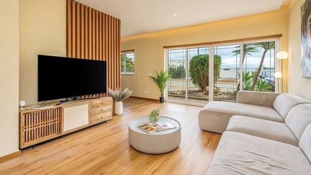 A modern living room with a flat-screen TV, a round coffee table, a beige sofa, and a large window offering a view of palm trees and a beach. - Deluxe House | Terrace & BBC by HostWise (Parchal)