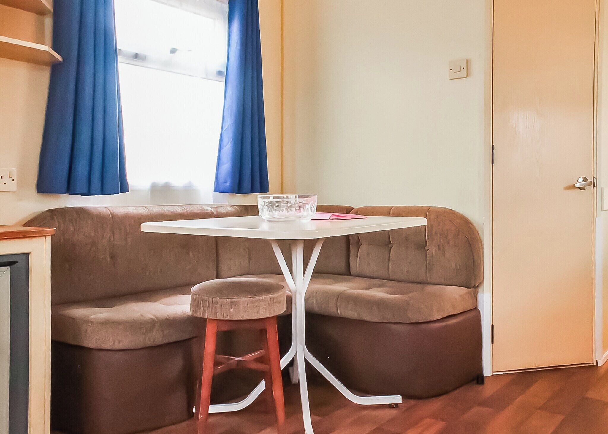 Un coin cuisine avec un plan de travail, une table et des tabourets blancs, une fenêtre avec des rideaux bleus et une porte.