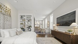 A modern bedroom with a large bed, a built-in wardrobe, and a dining area in the background.