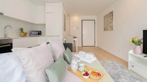 Ein modernes, helles Zimmer mit einem Bett, einer kleinen Küchenzeile, einem Essbereich mit Obstplatte und einem Fernseher auf einem weißen Schrank.