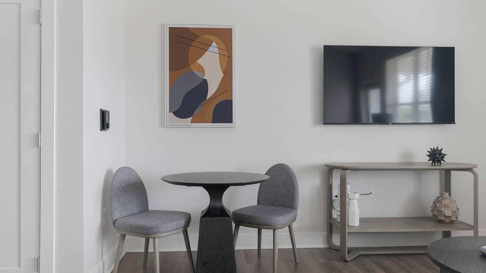 A modern living room with a round table and two grey upholstered chairs, a flat-screen TV mounted on the wall, and a minimalist console table with a decorative item.