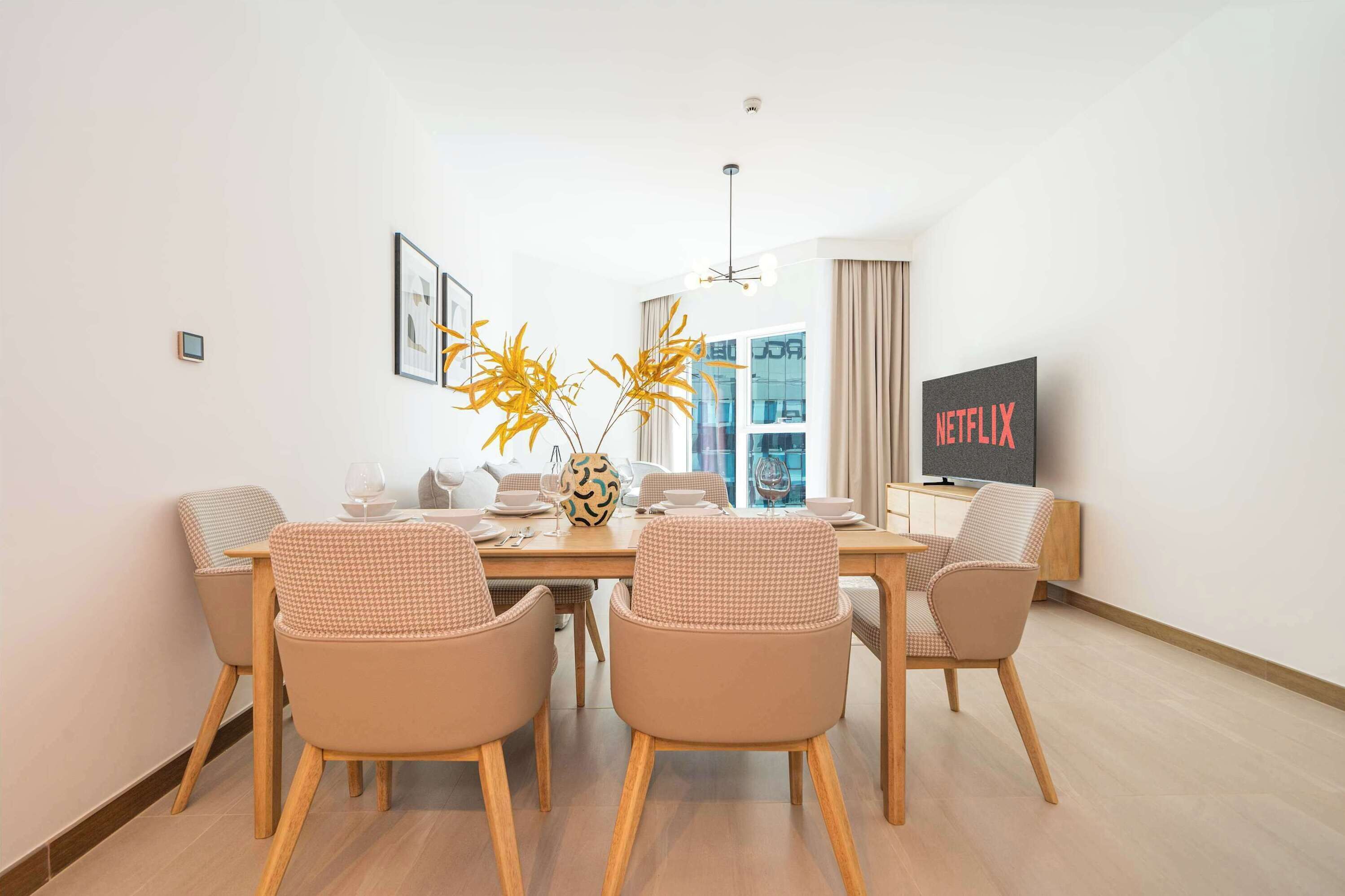 Un espace repas moderne avec une table en bois, des chaises de couleur beige et un vase rempli de fleurs jaunes. On peut apercevoir un téléviseur affichant le logo « NETFLIX » à l’arrière-plan.