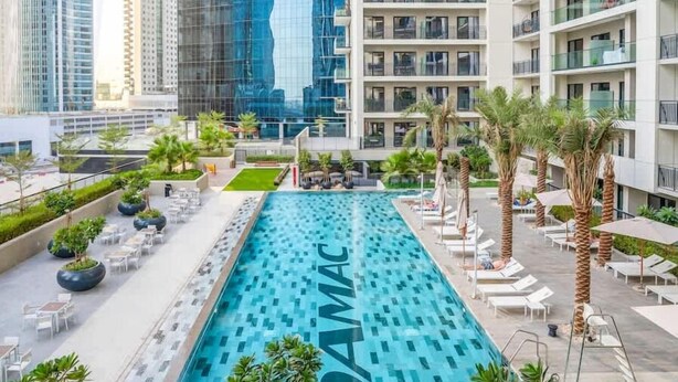 A rooftop swimming pool with a modern design, surrounded by lounge areas and palm trees. - Spacious & Fully Furnished 1BR in Zada (Dubai)