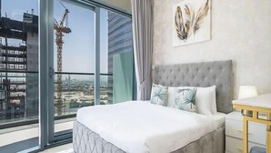 A bedroom with a large bed, a nightstand, a lamp, and a view of a construction site through a large window. - Spacious & Fully Furnished 1BR in Zada (Dubai)