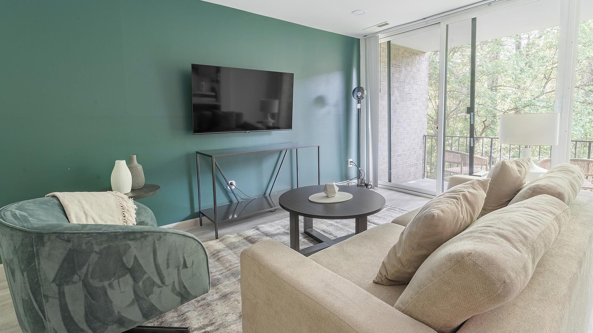 Una sala moderna con una pared color turquesa, un televisor de pantalla plana, una mesa de centro redonda, un sofá y un balcón con vistas a árboles.