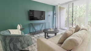 A modern living room with a teal wall, a flat-screen TV, a round coffee table, a sofa, and a balcony with a view of trees.