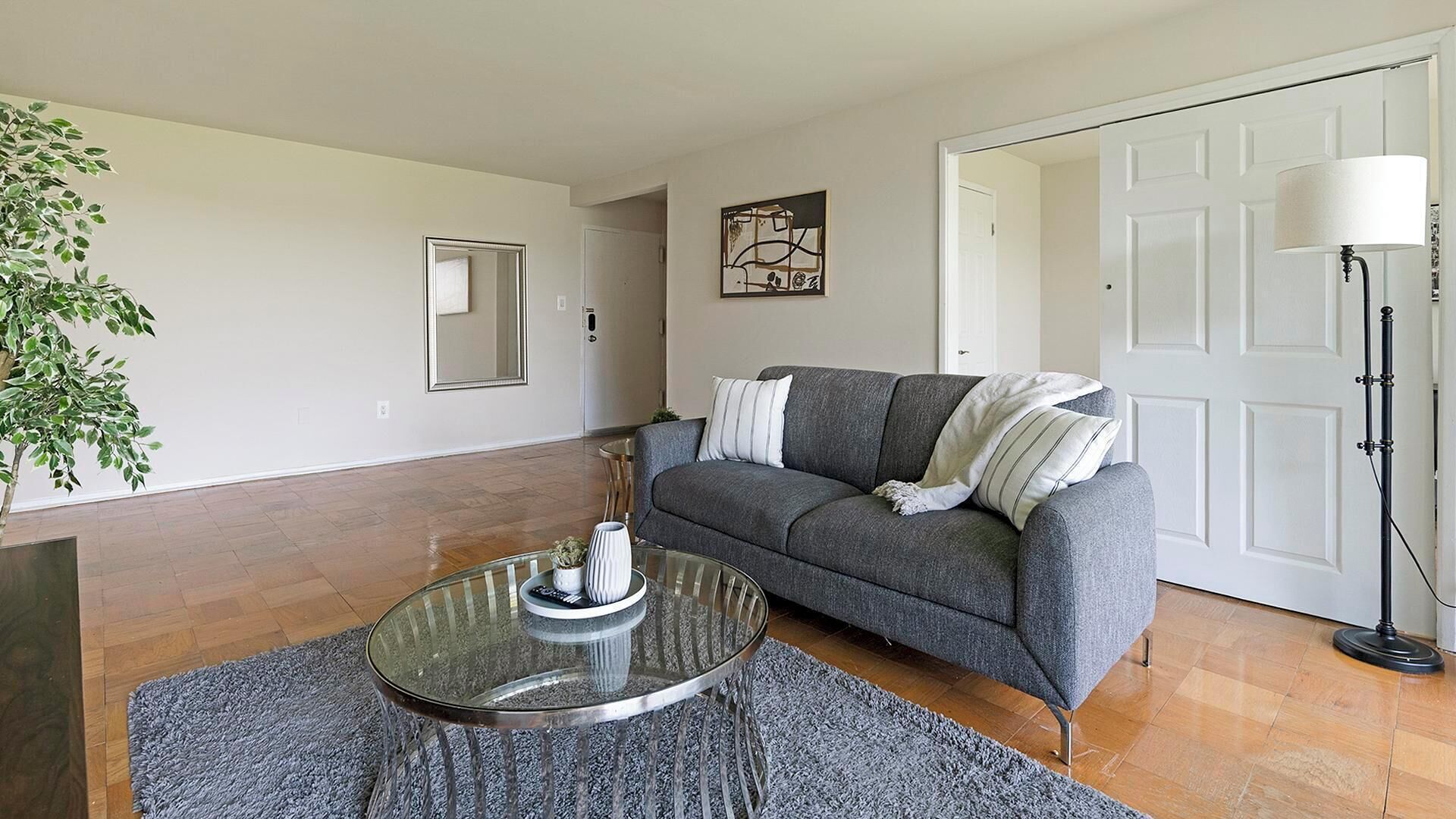 A living room with a grey sofa, a glass coffee table, a plant, and a painting on the wall.