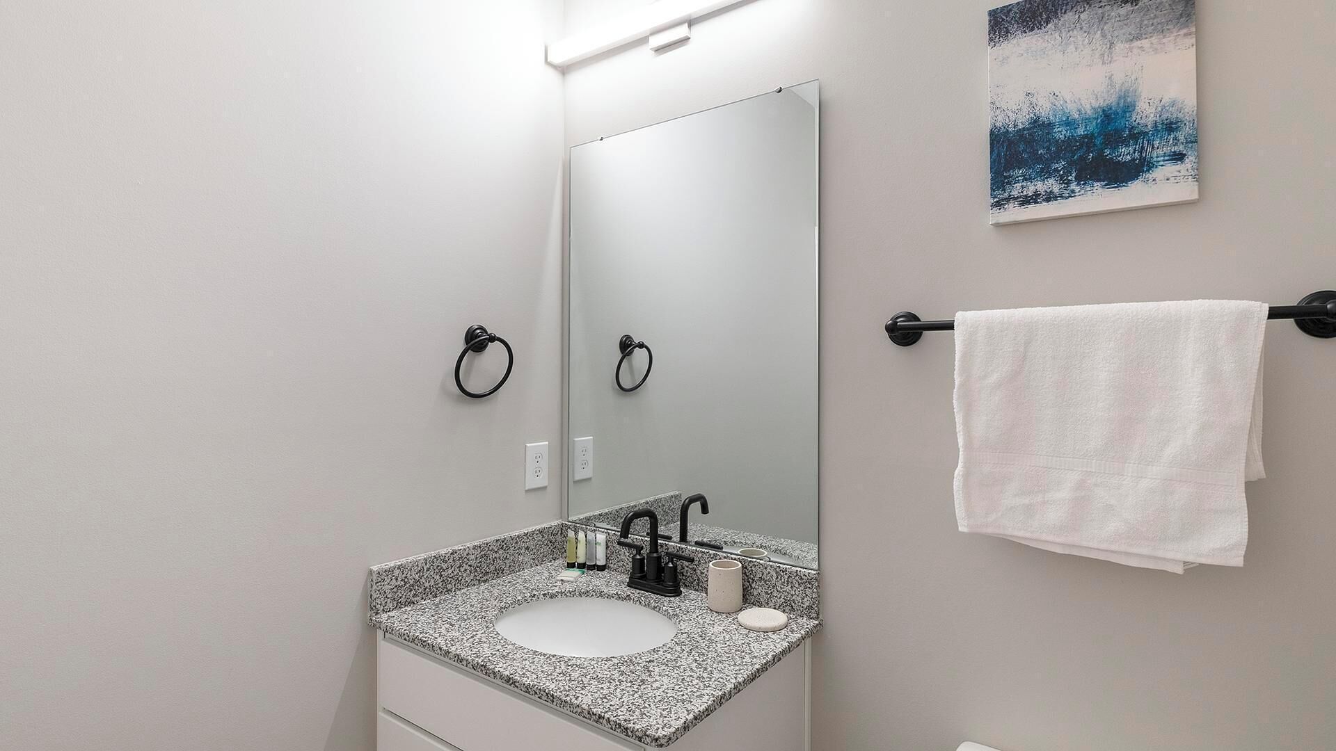 Un baño con lavamanos de granito, espejo, toallero y un cuadro en la pared.