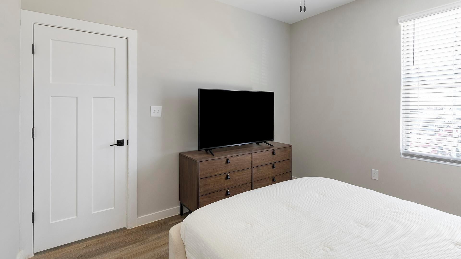 A bedroom with a white door, a TV on a wooden dresser, a bed with a white mattress, and a window with blinds.