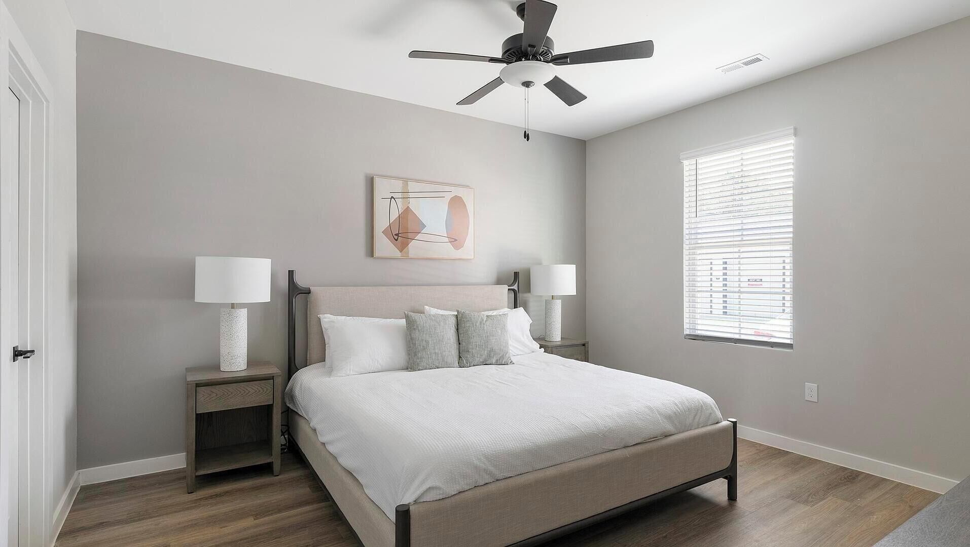 A bedroom with a bed, nightstands, a ceiling fan, a window with blinds, and a wall-mounted artwork.