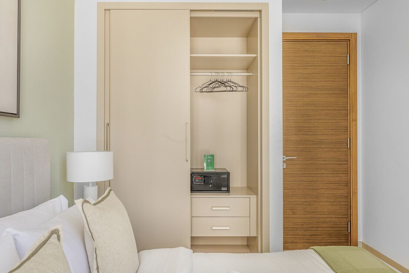 Una camera d'albergo con un letto, un comodino con una lampada, un armadio con grucce, una piccola cassettiera e una porta in legno.