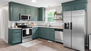 A modern kitchen with teal cabinets, stainless steel appliances, and a window with a view of greenery.