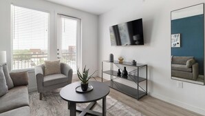 Una sala de estar moderna con un sofá gris, una mesa redonda con una planta, una consola de vidrio con adornos, un televisor montado y un espejo grande en la pared.