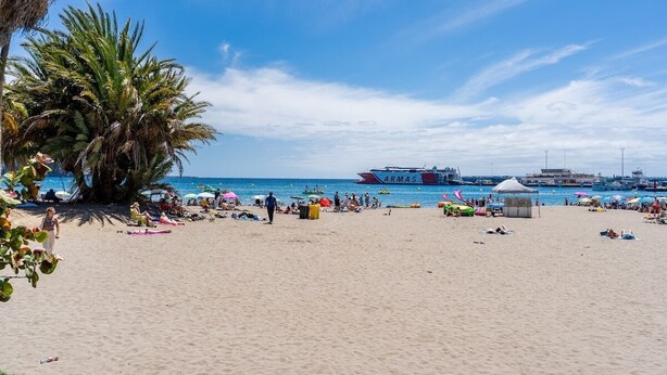Ein Strand mit Palmen, Sonnenschirmen und entspannenden Menschen. Im Hintergrund ist ein großes Schiff mit der Aufschrift 'ARMAS' zu sehen.