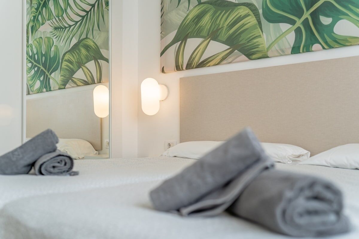A neatly made bed with rolled towels, a wall-mounted light, and a large tropical leaf-patterned painting.