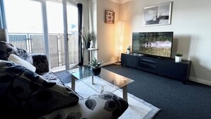 A modern living room with a sofa, a glass coffee table, a TV on a black stand, and a balcony with a view.