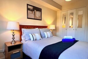 Un lit bien fait, recouvert d’une couverture sombre, avec deux lampes de chevet et une table de chevet sur laquelle se trouve une lampe.