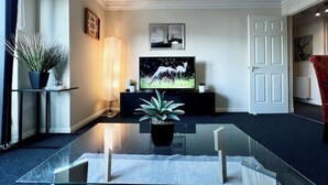 A modern living room with a glass coffee table, a flat-screen TV, a potted plant, and a framed picture on the wall.