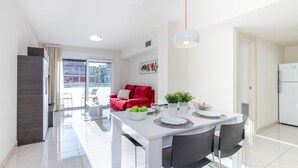 A modern kitchen with a white refrigerator, a dining table set with plates and a bowl of fruit, and a red sofa in the living area.